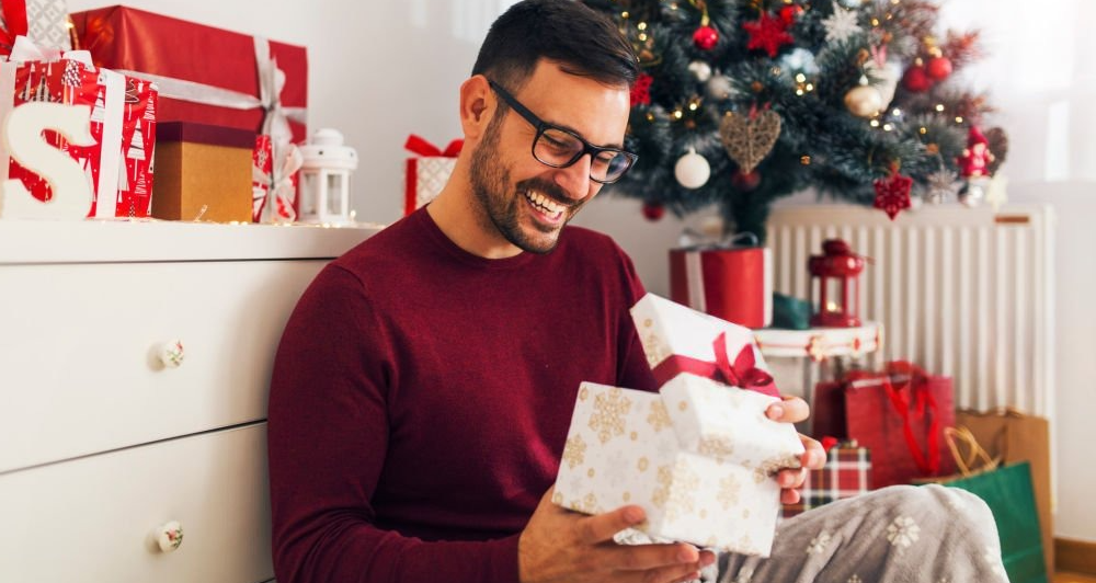 Man excitedly opening a Christmas gift with a smile, surrounded by holiday decorations and festive atmosphere